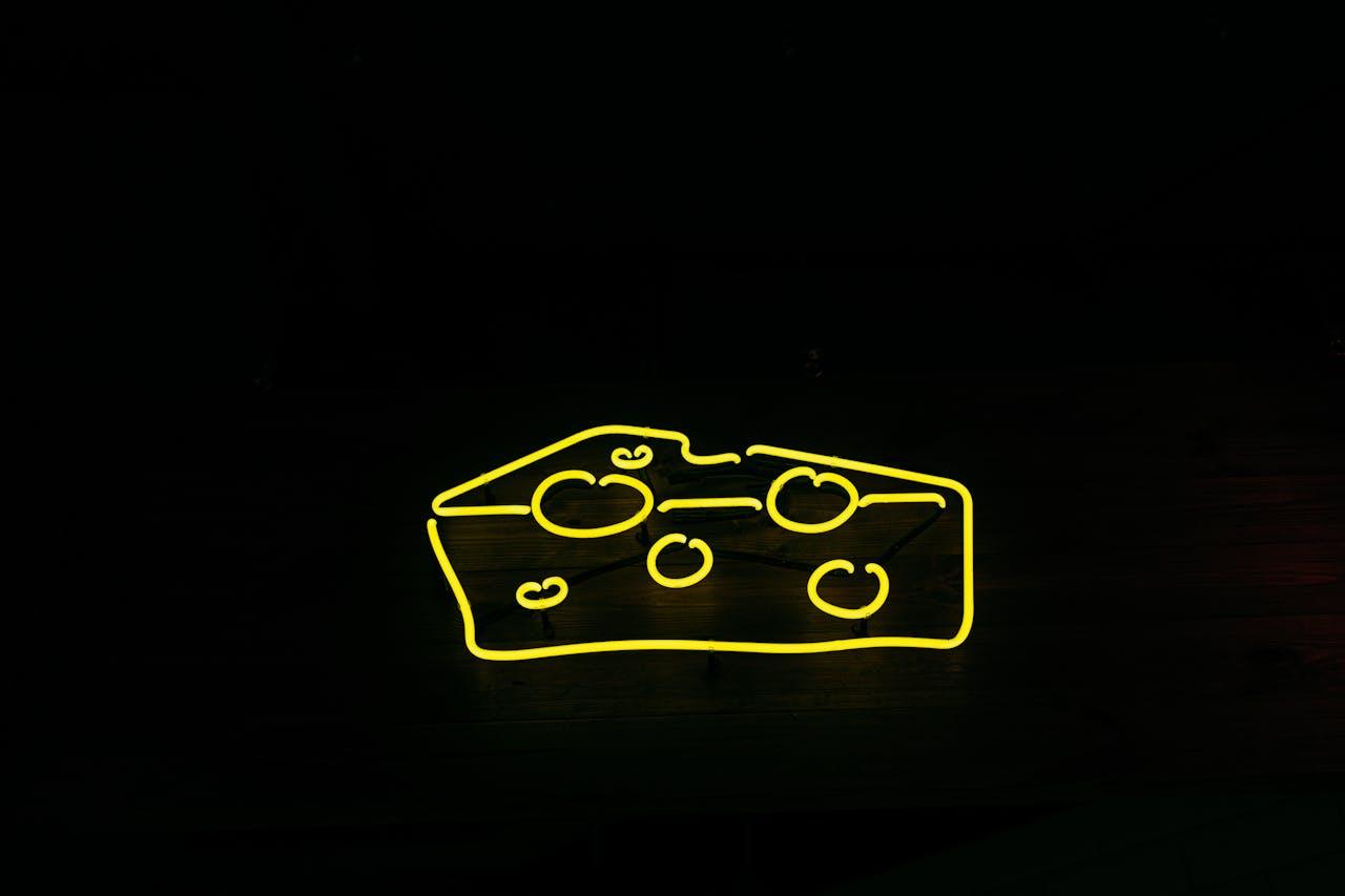 A bright yellow neon sign shaped like cheese against a black background, creating a modern and eye-catching design.